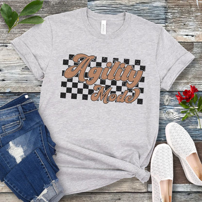 A grey dog agility t-shirt with 'Agility Mode' written in a retro stylized font over a checkerboard pattern, placed on a wooden surface, accompanied by jeans and white shoes.