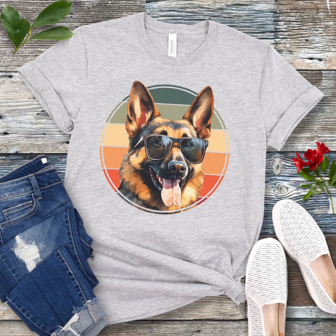 Grey t-shirt for German Shepherd lovers with smiling Shepherd dog wearing sunglasses and vintage sunset graphic, shown on a wooden surface with jeans and sneakers.