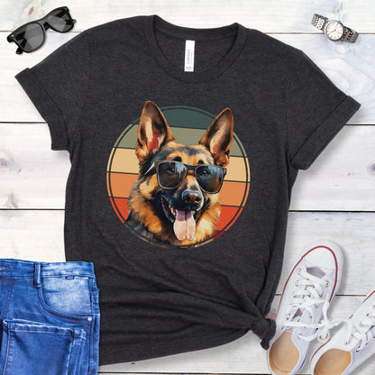 Dark grey t-shirt for German Shepherd lovers with smiling Shepherd dog wearing sunglasses and vintage sunset graphic, shown on a wooden surface with jeans and sneakers.
