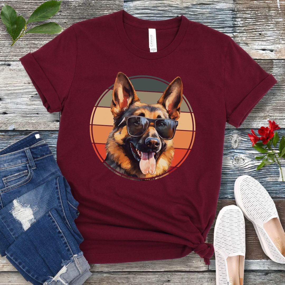 Maroon t-shirt for German Shepherd lovers with smiling Shepherd dog wearing sunglasses and vintage sunset graphic, shown on a wooden surface with jeans and sneakers.