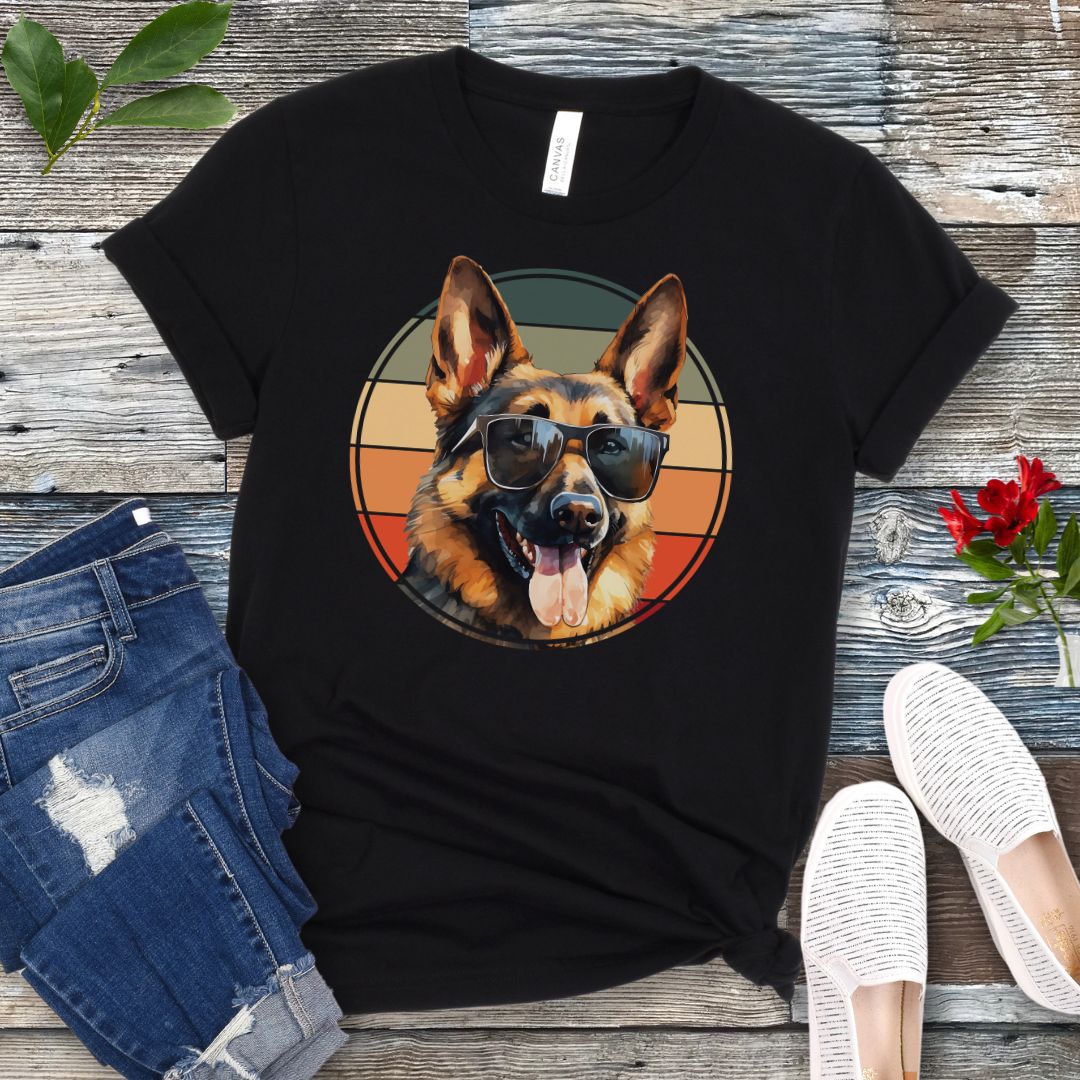 Black t-shirt for German Shepherd lovers with smiling Shepherd dog wearing sunglasses and vintage sunset graphic, shown on a wooden surface with jeans and sneakers.