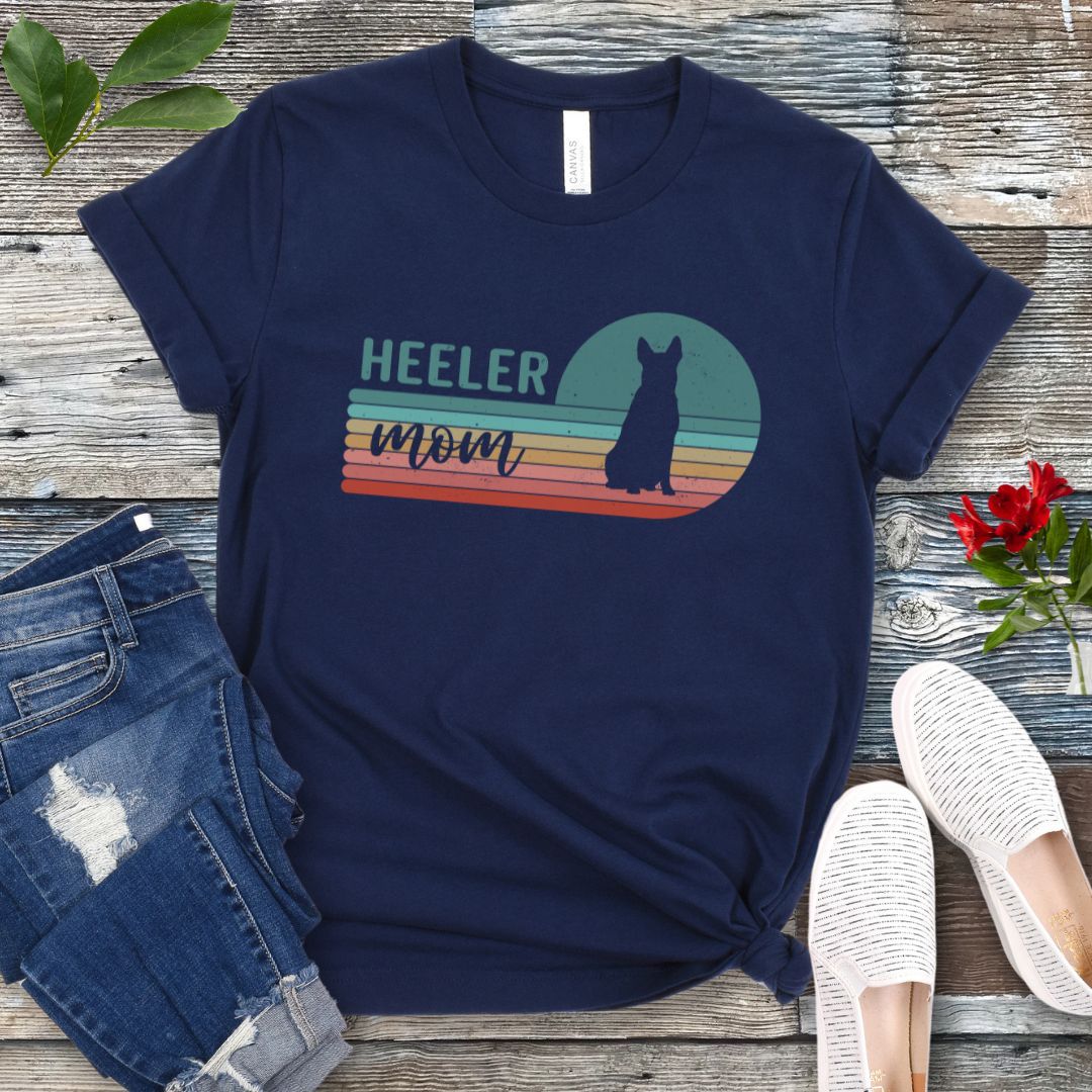 A navy Australian Cattle Dog t-shirt with retro sunset stripes, the text 'HEELER mom' and a Heeler silhouette in the center, placed on a wooden surface with a pair of jeans and white shoes in the background.