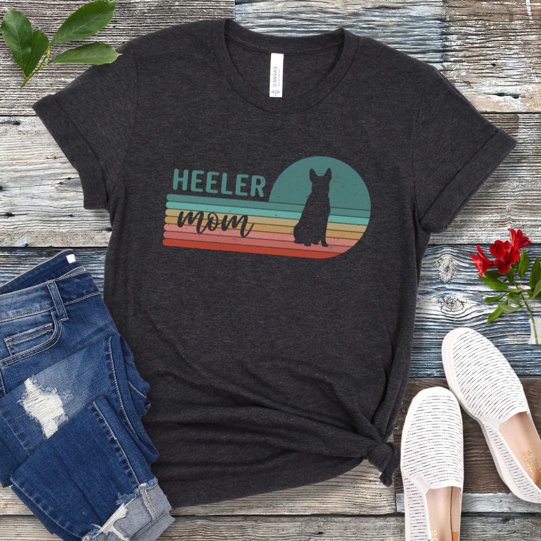 A dark grey Australian Cattle Dog t-shirt with retro sunset stripes, the text 'HEELER mom' and a Heeler silhouette in the center, placed on a wooden surface with a pair of jeans and white shoes in the background.
