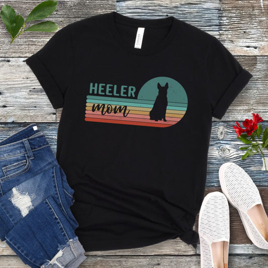 A black Australian Cattle Dog t-shirt with retro sunset stripes, the text 'HEELER mom' and a Heeler silhouette in the center, placed on a wooden surface with a pair of jeans and white shoes in the background.
