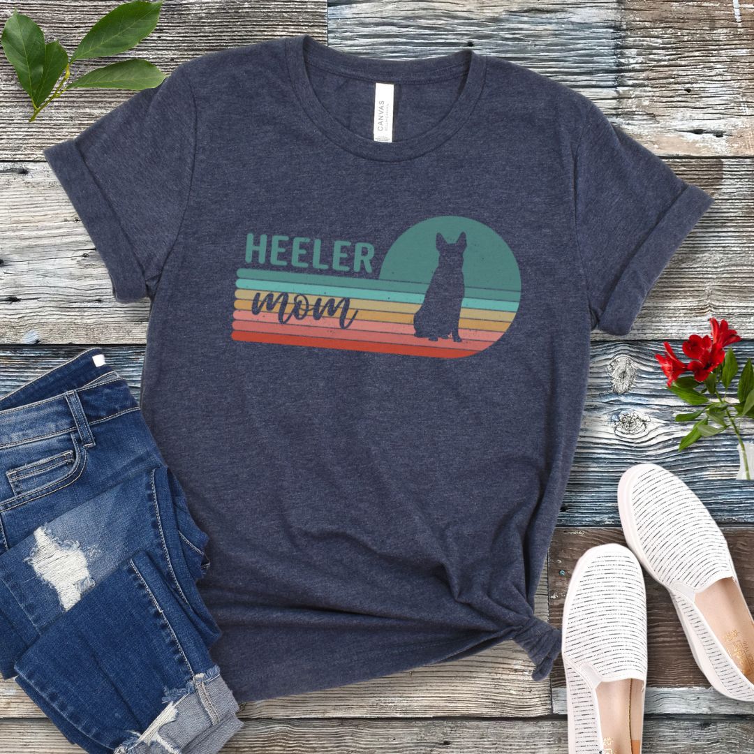 A heather navy Australian Cattle Dog t-shirt with retro sunset stripes, the text 'HEELER mom' and a Heeler silhouette in the center, placed on a wooden surface with a pair of jeans and white shoes in the background.