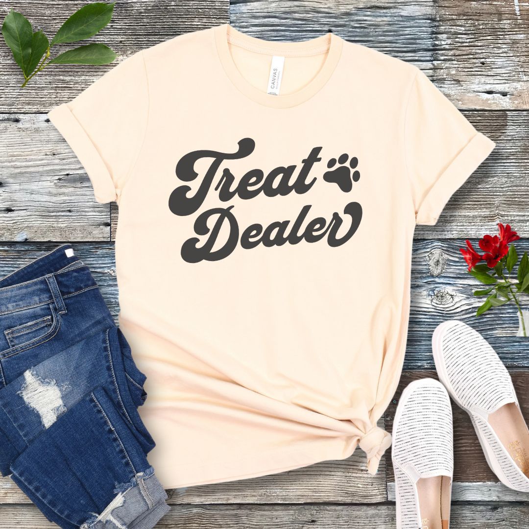 A beige T-shirt with the retro font with the text 'Treat Dealer' printed on the front, accompanied by a paw print. The shirt is displayed on a wooden background with a pair of jeans and white shoes.