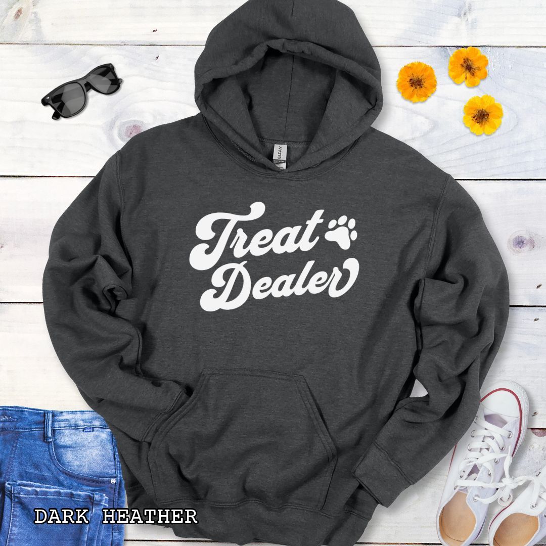 Dark grey hoodie with the text 'Treat Dealer' printed on the front in retro font, accompanied by a paw print. The shirt is displayed on a wooden background with a pair of jeans and white shoes.