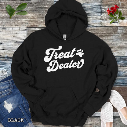 Black hoodie with the text 'Treat Dealer' printed on the front in retro font, accompanied by a paw print. The shirt is displayed on a wooden background with a pair of jeans and white shoes.