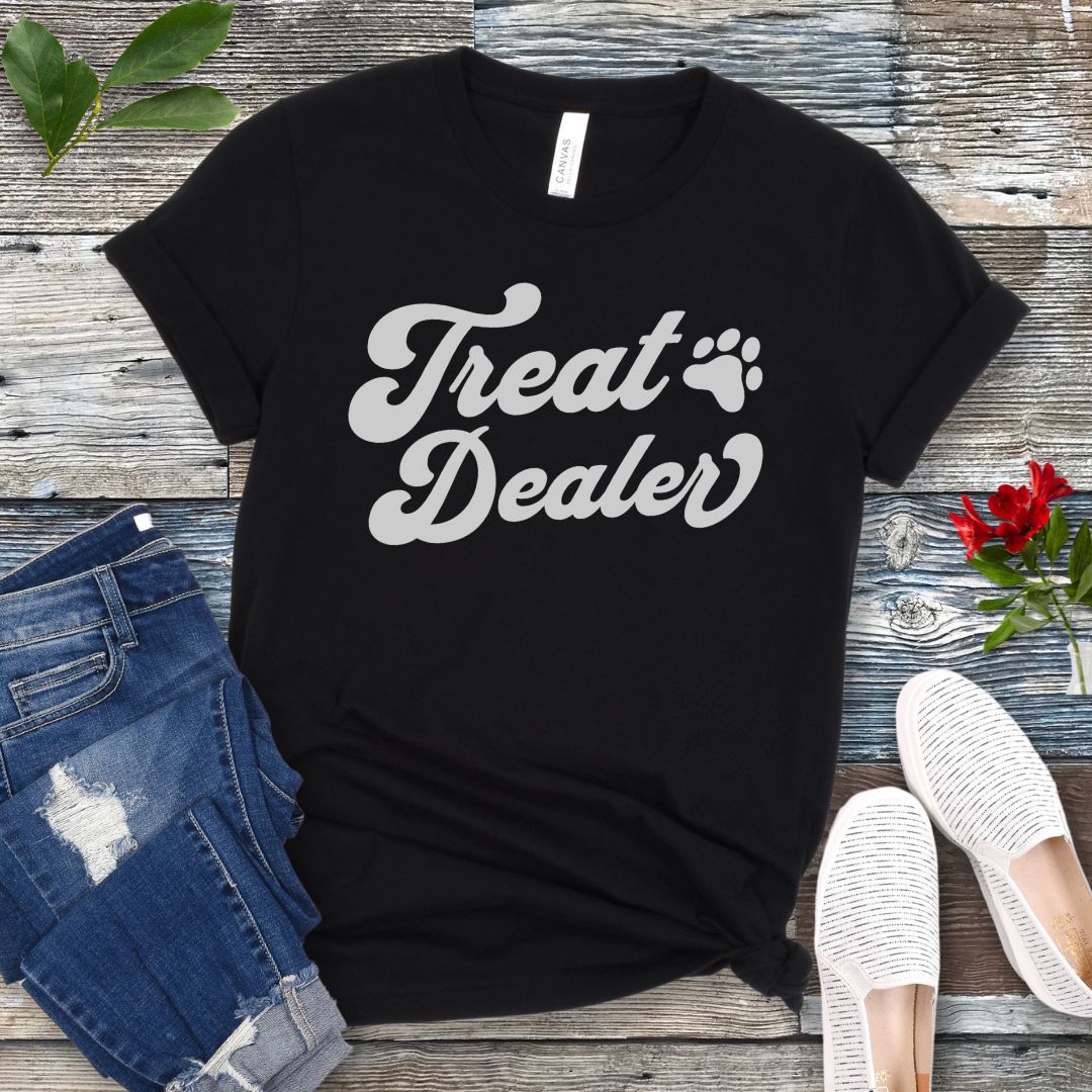 A black T-shirt with the retro font with the text 'Treat Dealer' printed on the front, accompanied by a paw print. The shirt is displayed on a wooden background with a pair of jeans and white shoes.