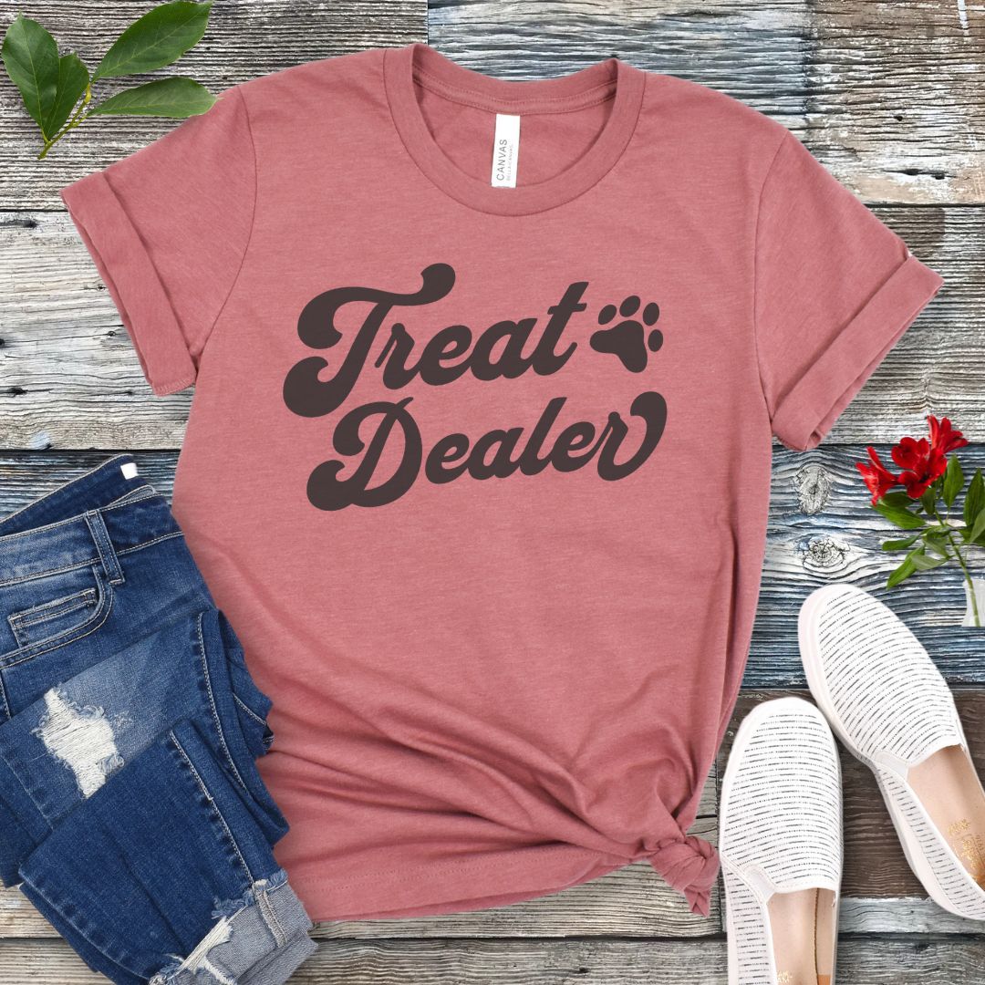 A mauve T-shirt with the retro font with the text 'Treat Dealer' printed on the front, accompanied by a paw print. The shirt is displayed on a wooden background with a pair of jeans and white shoes.
