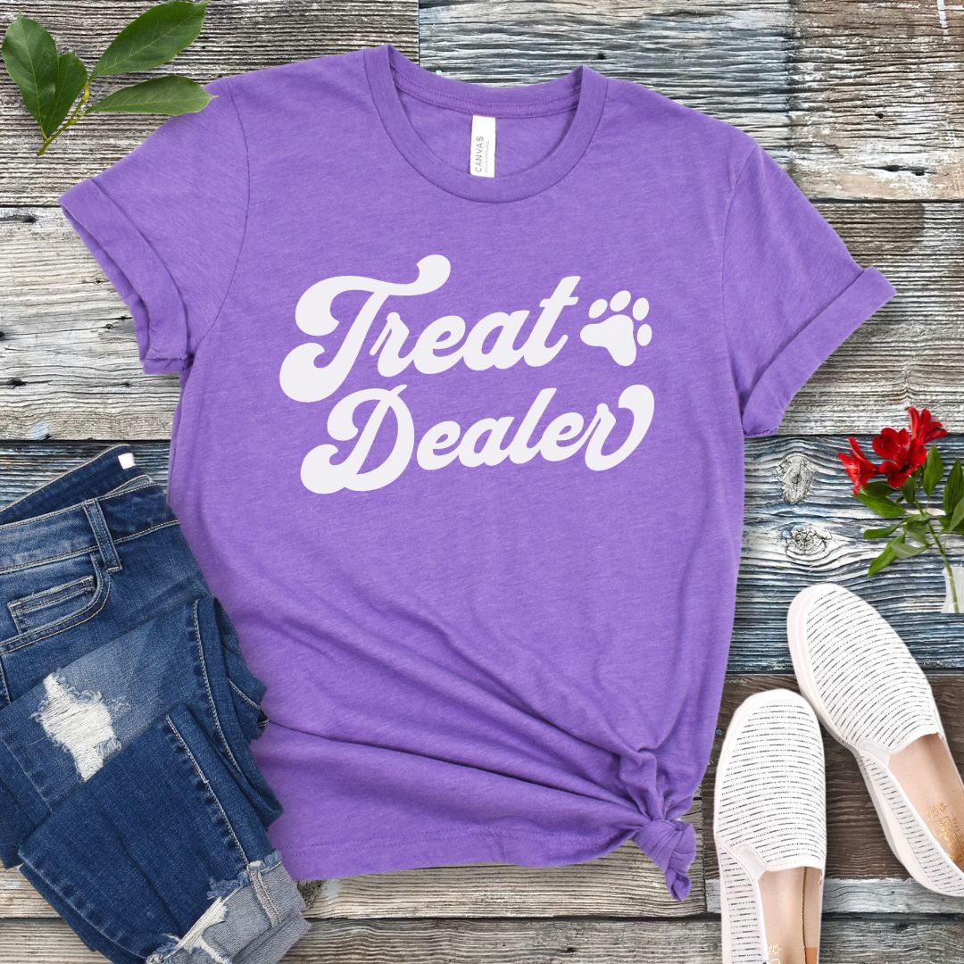 A purple T-shirt with the retro font with the text 'Treat Dealer' printed on the front, accompanied by a paw print. The shirt is displayed on a wooden background with a pair of jeans and white shoes.