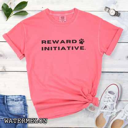 Pink (Watermelon) Comfort Colors dog training t-shirt with 'REWARD INITIATIVE' text, presented on a wooden surface with jeans, sneakers, and sunglasses.