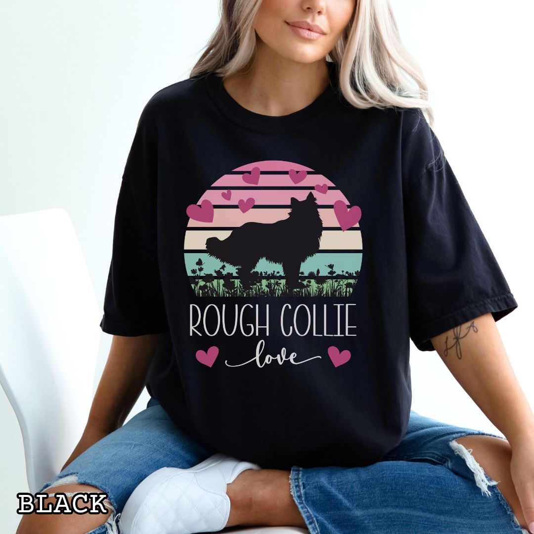Person wearing a black Comfort Colors t-shirt with colorful vintage sunset design with hearts, flowers, and Rough Collie silhouette and the text 'Rough Collie Love', shown on a wooden surface.