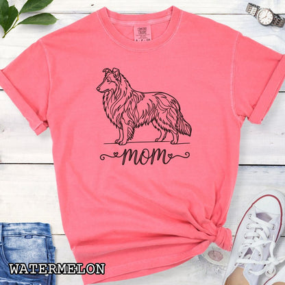 Pink Comfort Colors t-shirt with a line art Rough Collie dog illustration and 'mom' text in feminine script beneath, shown on a wooden surface with denim jeans and a flower.