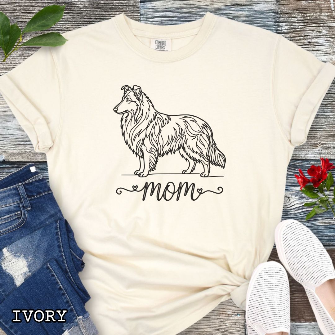Beige Comfort Colors t-shirt with a line art Rough Collie dog illustration and 'mom' text in feminine script beneath, shown on a wooden surface with denim jeans and a flower.