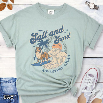 Bay Comfort Colors t-shirt for Rough Collie lovers with retro surfing Collie dog graphic in a beach setting with 'Salt and Sand Adventure Squad' text, shown on a wooden surface.