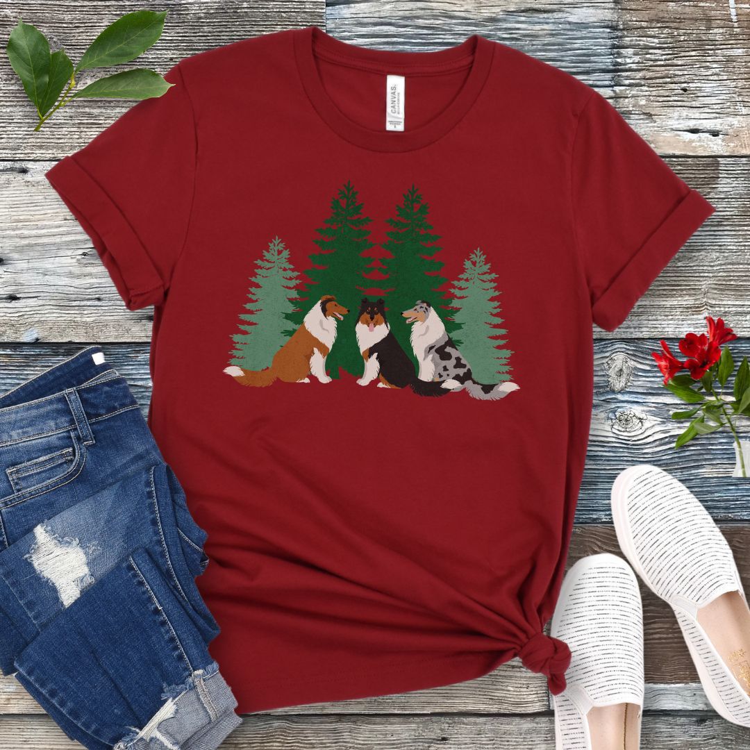 Cardinal red Collie lover t-shirt with tricolor, sable and white and blue merle Rough Collies sitting in front of evergreen trees, shown on a wooden surface with jeans and shoes.