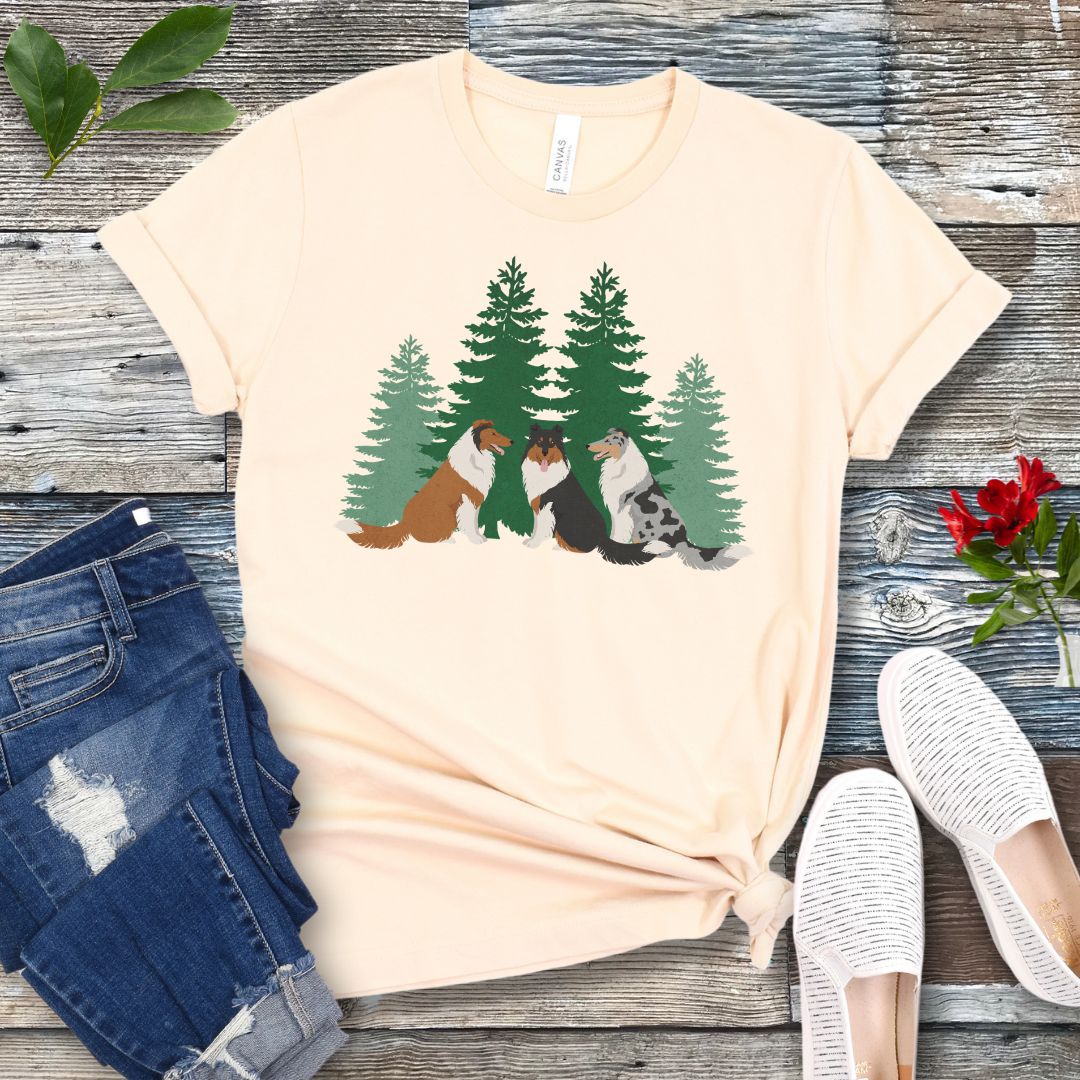 Beige Collie lover t-shirt with tricolor, sable and white and blue merle Rough Collies sitting in front of evergreen trees, shown on a wooden surface with jeans and shoes.