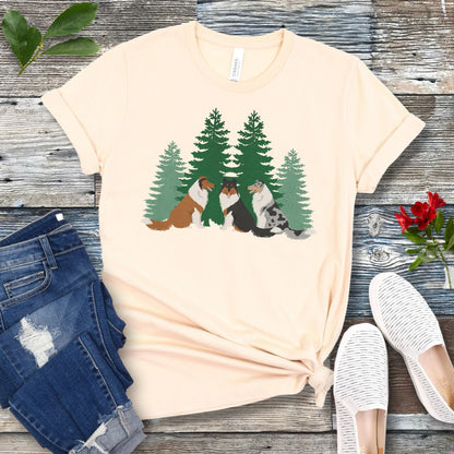 Beige Collie lover t-shirt with tricolor, sable and white and blue merle Rough Collies sitting in front of evergreen trees, shown on a wooden surface with jeans and shoes.