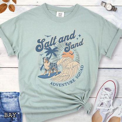 Pale green Comfort Colors t-shirt for Australian Shepherd lovers with beach-themed graphic of a surfing Aussie dog, waves and sunset, and the text 'Salt and Sand Adventure Squad' in retro lettering, shown on a wooden surface with accessories.