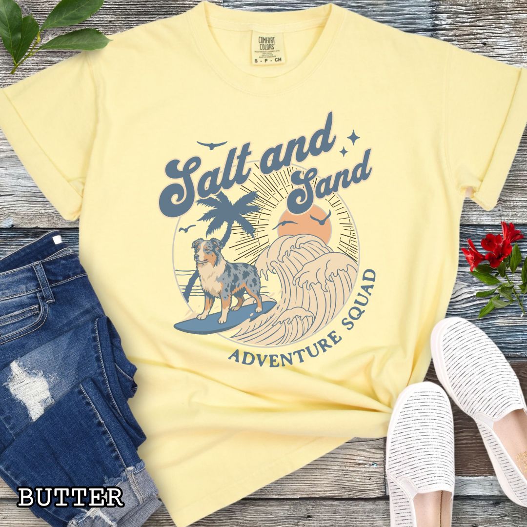 Yellow Comfort Colors t-shirt for Australian Shepherd lovers with beach-themed graphic of a surfing Aussie dog, waves and sunset, and the text 'Salt and Sand Adventure Squad' in retro lettering, shown on a wooden surface with accessories.