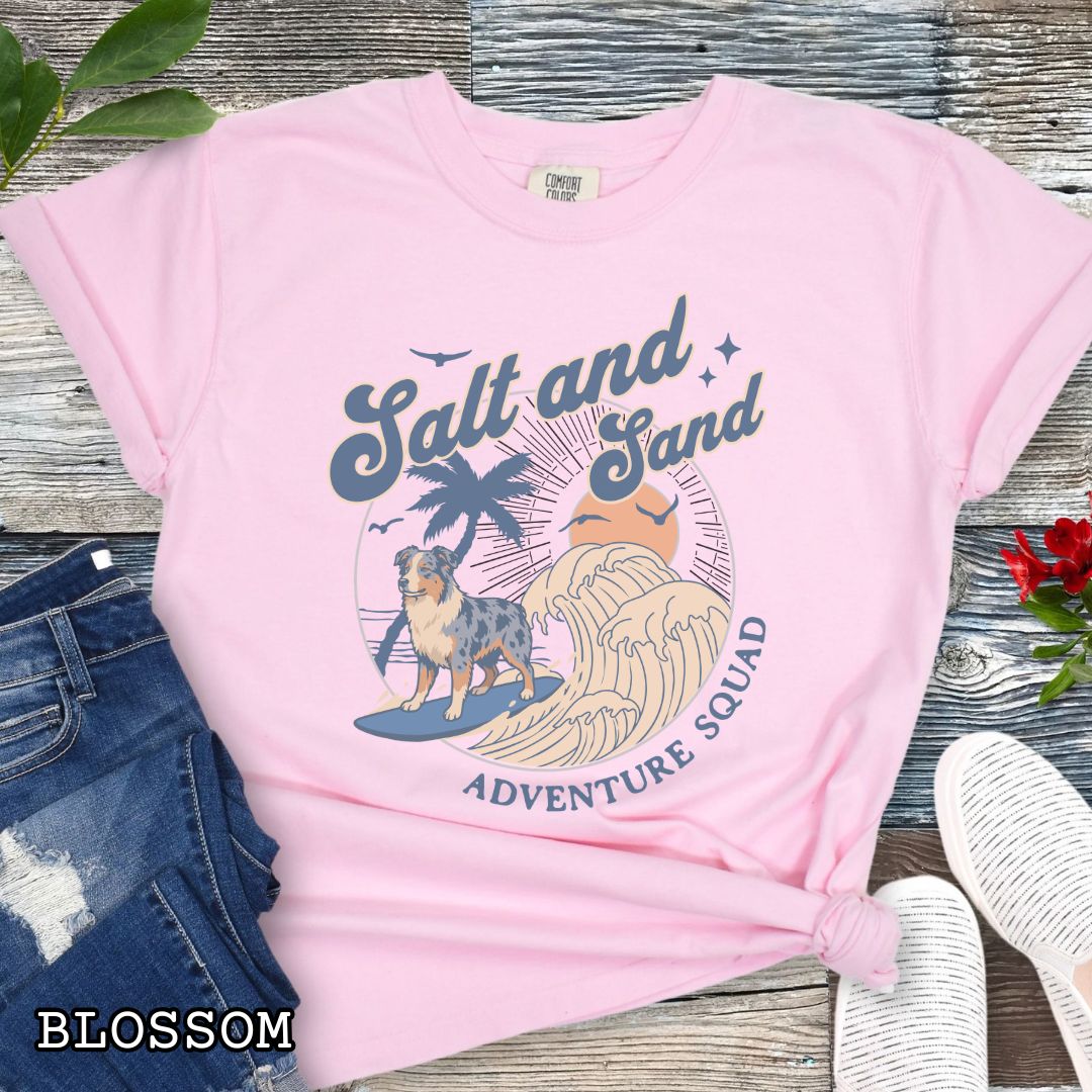 Pink Comfort Colors t-shirt for Australian Shepherd lovers with beach-themed graphic of a surfing Aussie dog, waves and sunset, and the text 'Salt and Sand Adventure Squad' in retro lettering, shown on a wooden surface with accessories.