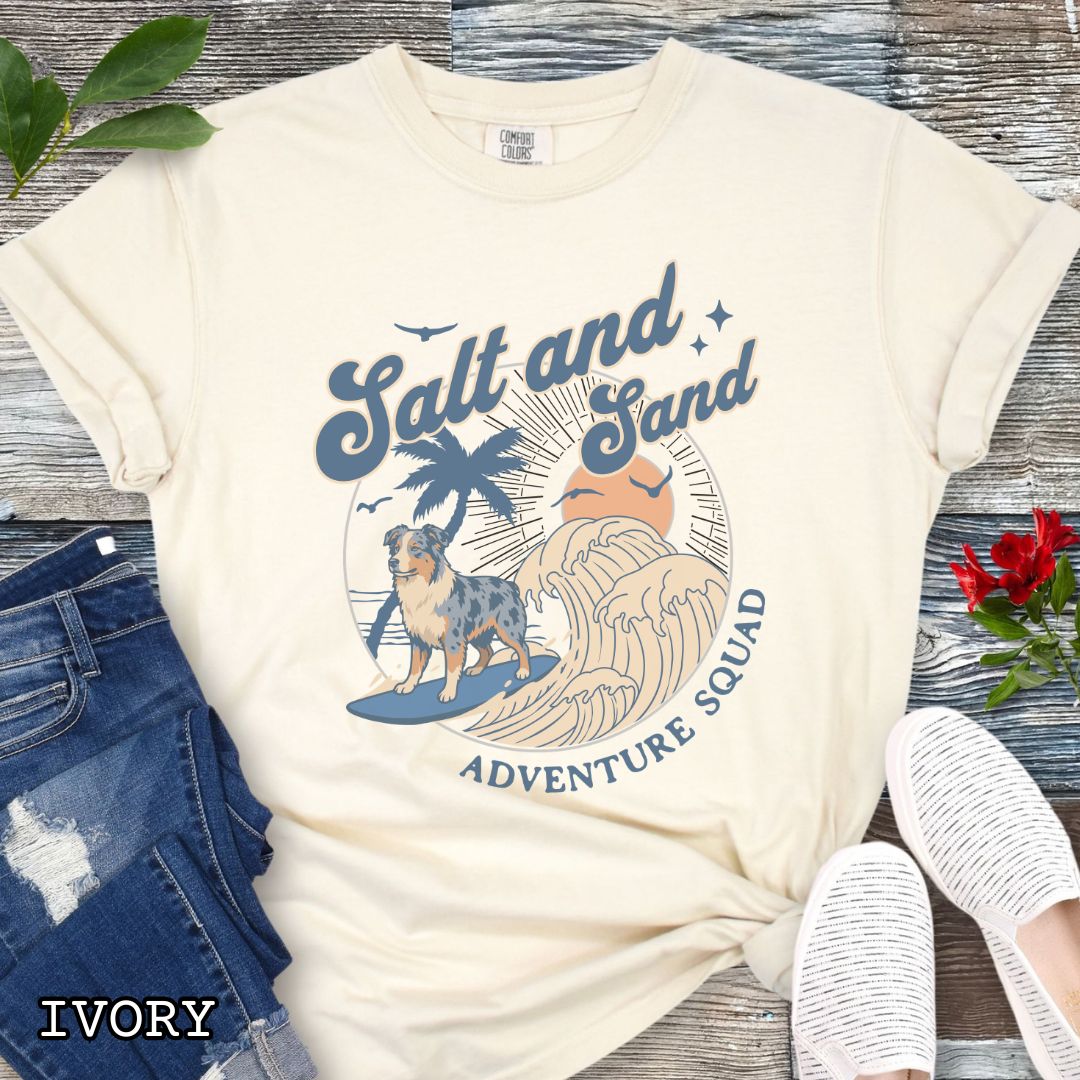Beige Comfort Colors t-shirt for Australian Shepherd lovers with beach-themed graphic of a surfing Aussie dog, waves and sunset, and the text 'Salt and Sand Adventure Squad' in retro lettering, shown on a wooden surface with accessories.
