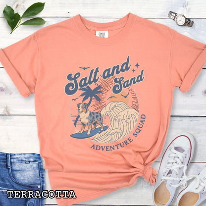 Orange Comfort Colors t-shirt for Australian Shepherd lovers with beach-themed graphic of a surfing Aussie dog, waves and sunset, and the text 'Salt and Sand Adventure Squad' in retro lettering, shown on a wooden surface with accessories.