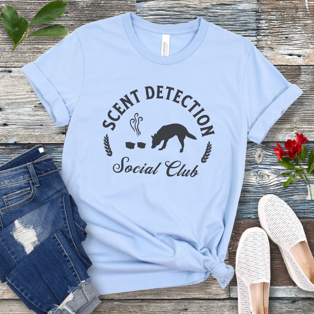 Light blue canine scent work t-shirt with 'Scent Detection Social Club' text and sniffing dog graphic, presented on a wooden surface with jeans and shoes.