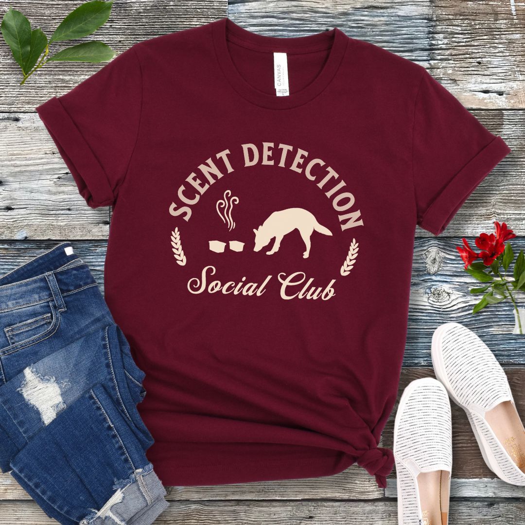 Maroon nosework t-shirt with 'Scent Detection Social Club' text and sniffing dog graphic, presented on a wooden surface with jeans and shoes.