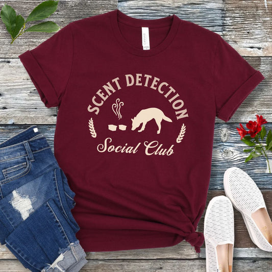Maroon nosework t-shirt with 'Scent Detection Social Club' text and sniffing dog graphic, presented on a wooden surface with jeans and shoes.