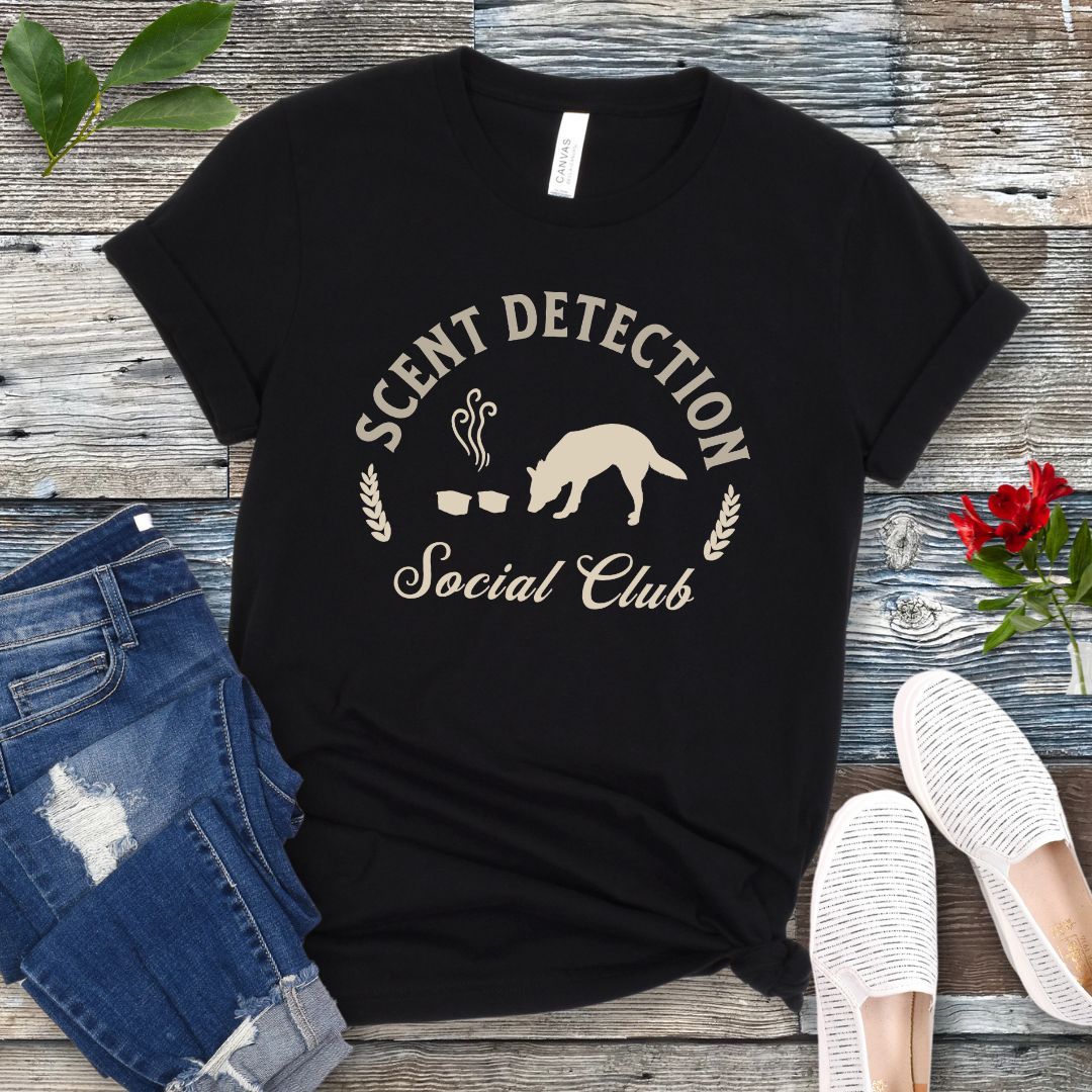 Black nosework t-shirt with 'Scent Detection Social Club' text and sniffing dog graphic, presented on a wooden surface with jeans and shoes.