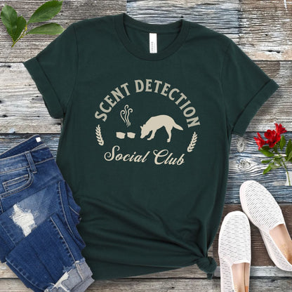 Dark green canine scent work t-shirt with 'Scent Detection Social Club' text and sniffing dog graphic, presented on a wooden surface with jeans and shoes.