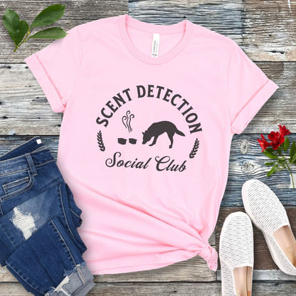 Pink canine scent work t-shirt with 'Scent Detection Social Club' text and sniffing dog graphic, presented on a wooden surface with jeans and shoes.
