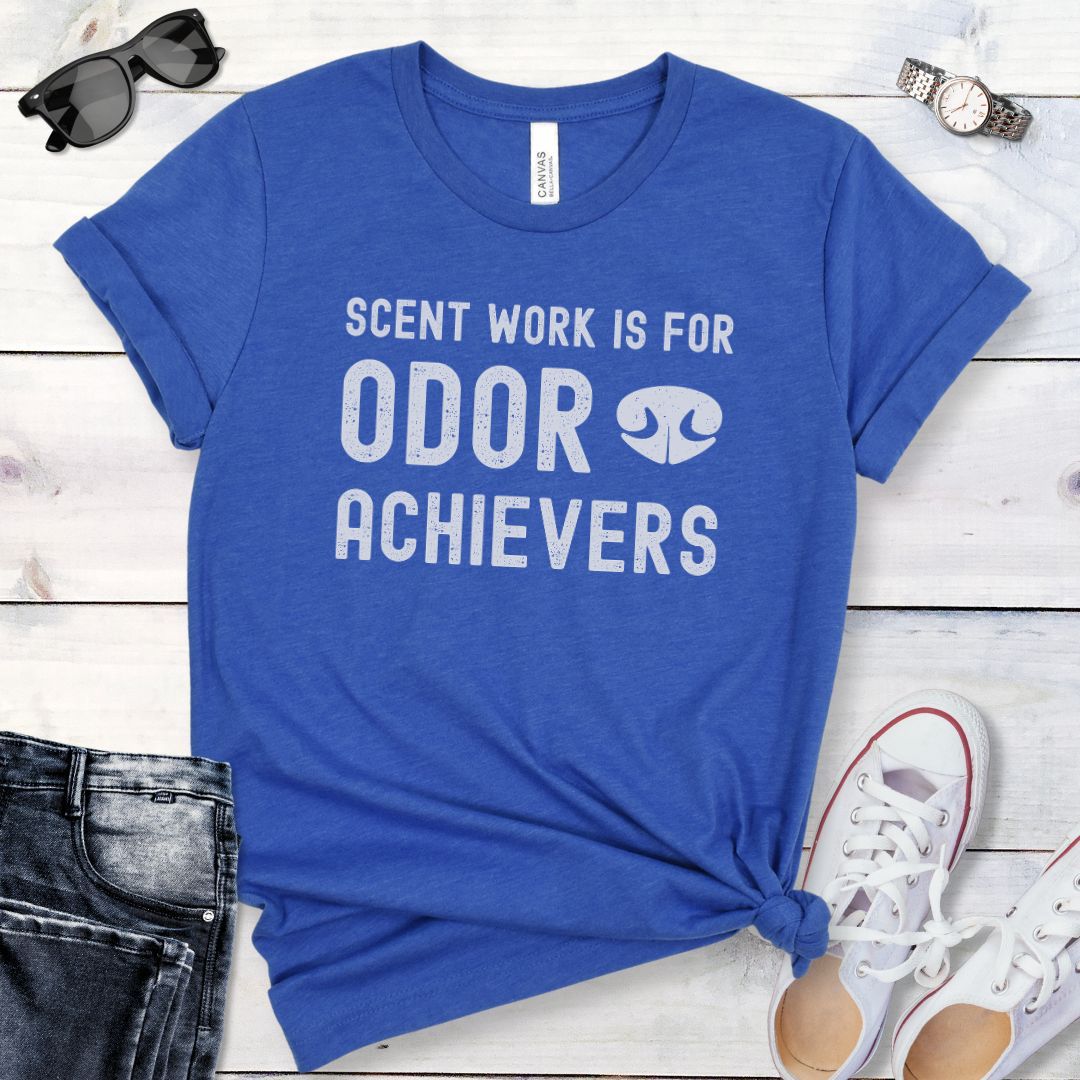 Royal blue t-shirt for dog scent detection trainers with humorous text 'Scent work is for odor achievers' and the graphic of a dog's nose, presented on a wooden surface with jeans and shoes.