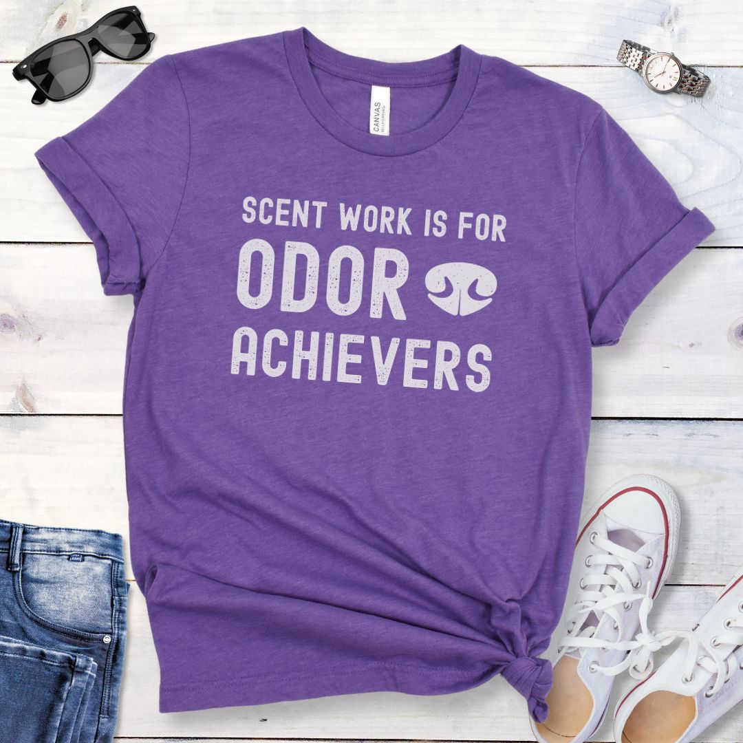 Purple t-shirt for dog scent detection trainers with humorous text 'Scent work is for odor achievers' and the graphic of a dog's nose, presented on a wooden surface with jeans and shoes.