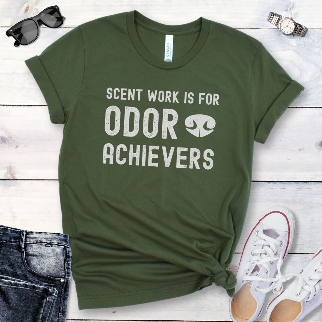 Military green t-shirt for dog scent detection trainers with humorous text 'Scent work is for odor achievers' and the graphic of a dog's nose, presented on a wooden surface with jeans and shoes.