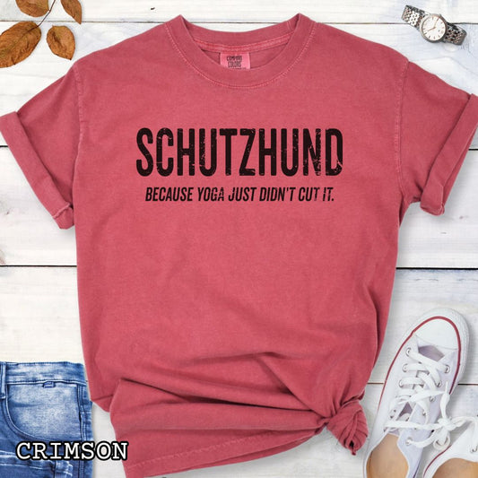 Red Comfort Colors t-shirt with 'Schutzhund because yoga just didn't cut it' text, shown on a wooden surface with jeans and sneakers.