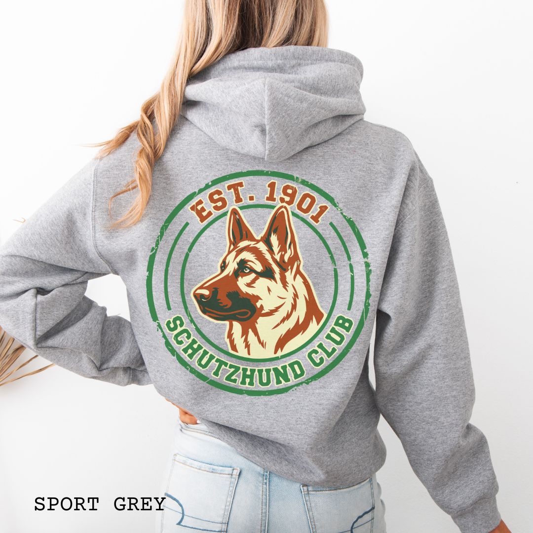 Grey back print hoodie with a  German Shepherd Dog and 'Established 1901, Schutzhund Club' text in a circular logo style design, displayed on a wooden surface with flowers and jeans.