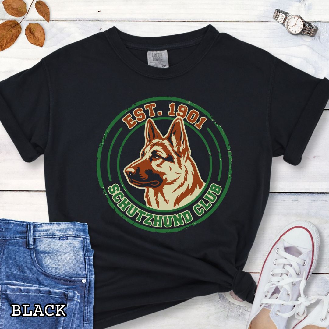 Black IGP dog sports t-shirt with a German Shepherd design and 'Est. 1901 Schutzhund Club' text on a wooden surface with jeans and sneakers.