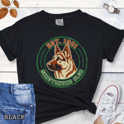 Black IGP dog sports t-shirt with a German Shepherd design and 'Est. 1901 Schutzhund Club' text on a wooden surface with jeans and sneakers.