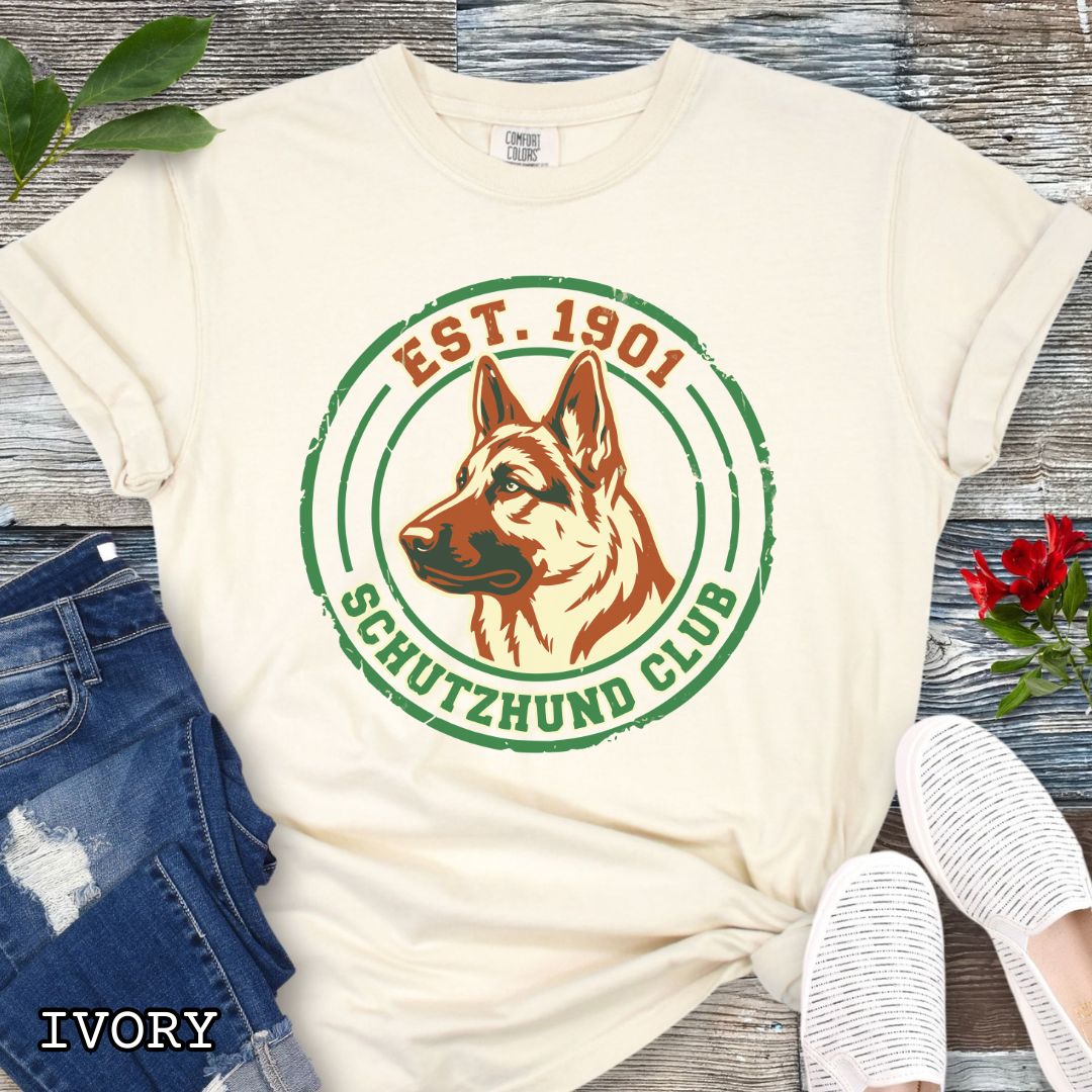 Beige IGP dog sports t-shirt with a German Shepherd design and 'Est. 1901 Schutzhund Club' text on a wooden surface with jeans and sneakers.