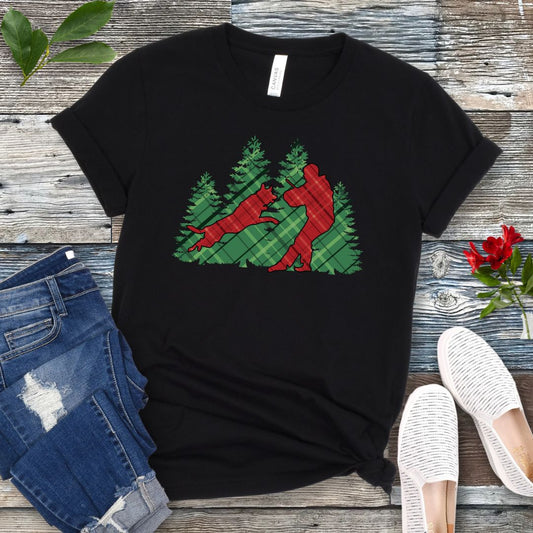 Black t-shirt with red and green plaid holiday graphic of a Schutzhund dog and handler doing bitework, presented on a wooden surface with jeans and shoes.