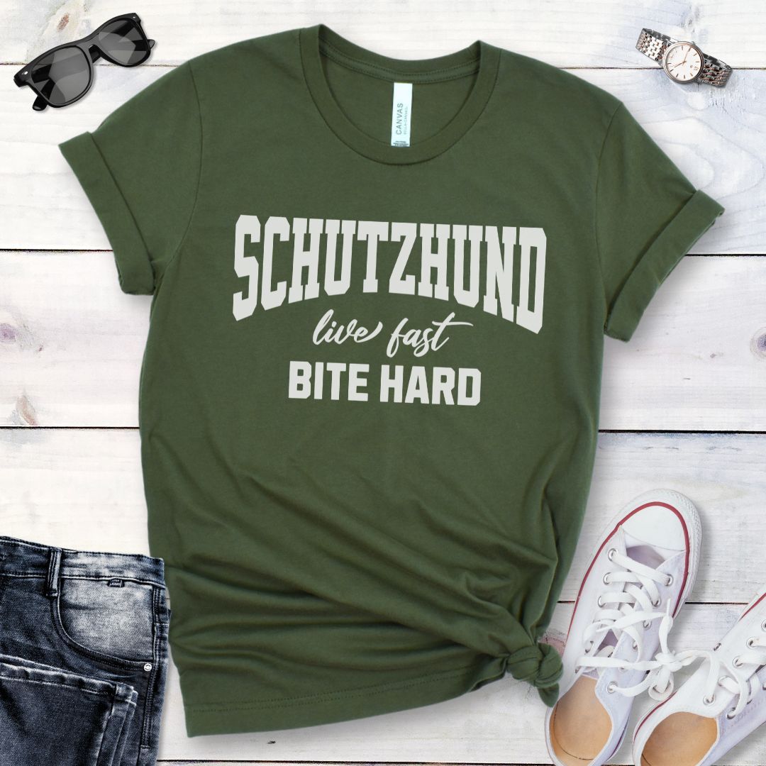 A military green bitework t-shirt with the text 'SCHUTZHUND live fast BITE HARD'  in bold, athletic style lettering. The shirt is displayed flat with a pair of blue rolled-up jeans and white shoes to the side, on a wooden surface with a red flower and green leaf partially visible.