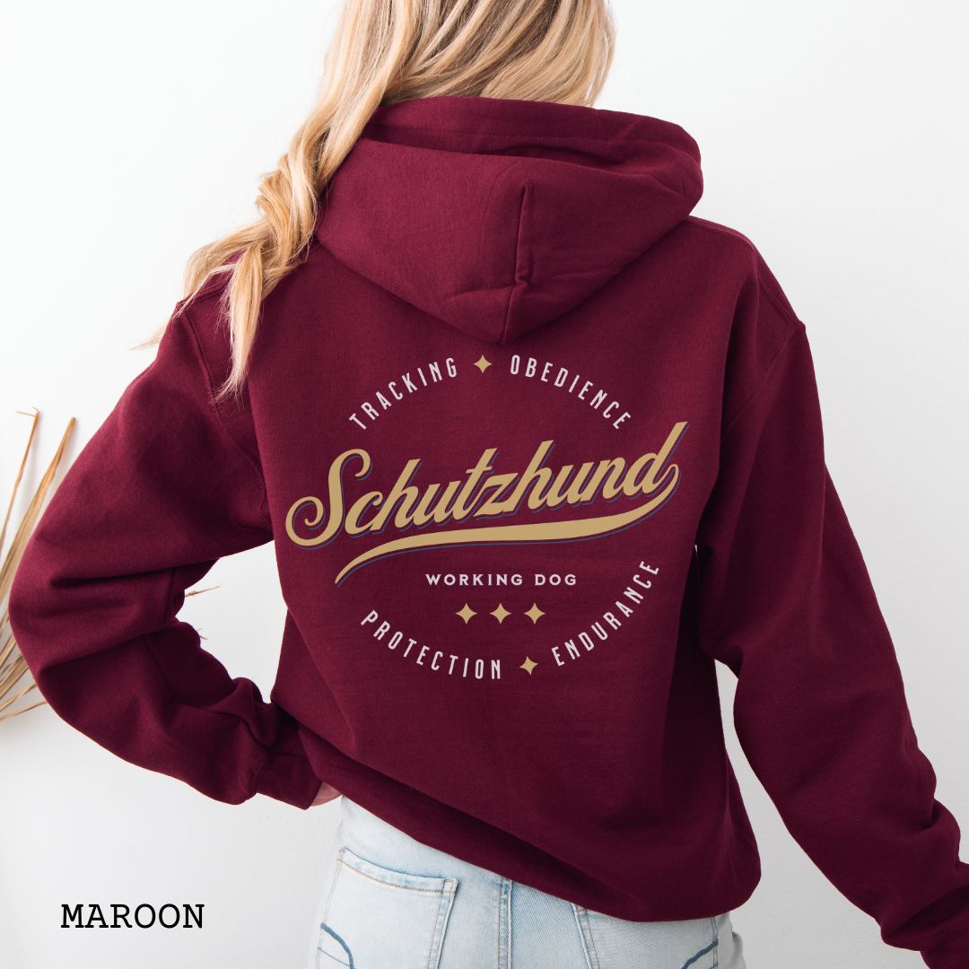 Back view of a maroon unisex Schutzhund/IGP hoodie with vintage style text design that reads 'Schutzhund, Working Dog, Tracking, Obedience, Protection, Endurance'. Presented on a wooden background with jeans and shoes.