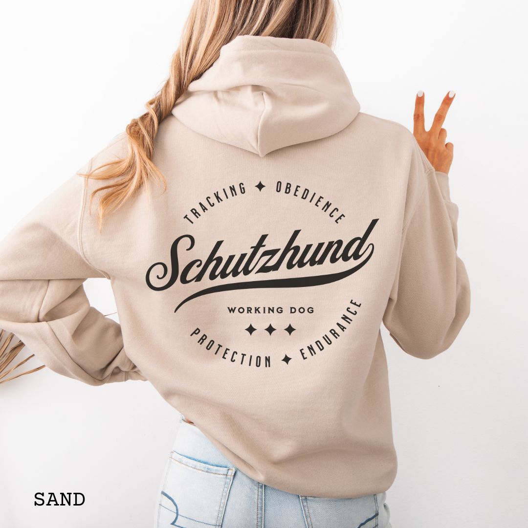 Back view of a beige unisex Schutzhund/IGP hoodie with vintage style text design that reads 'Schutzhund, Working Dog, Tracking, Obedience, Protection, Endurance'. Presented on a wooden background with jeans and shoes.