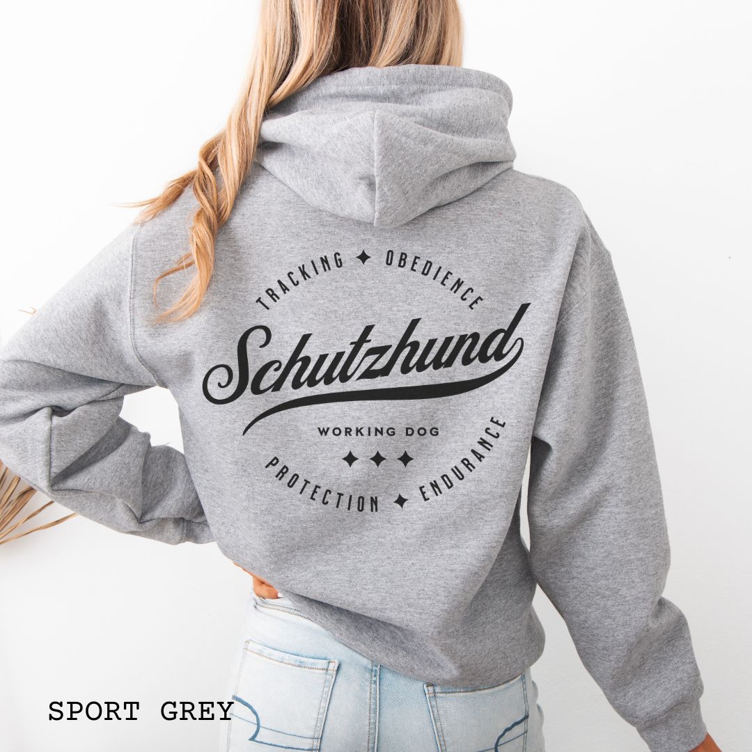 Back view of a grey unisex Schutzhund/IGP hoodie with vintage style text design that reads 'Schutzhund, Working Dog, Tracking, Obedience, Protection, Endurance'. Presented on a wooden background with jeans and shoes.