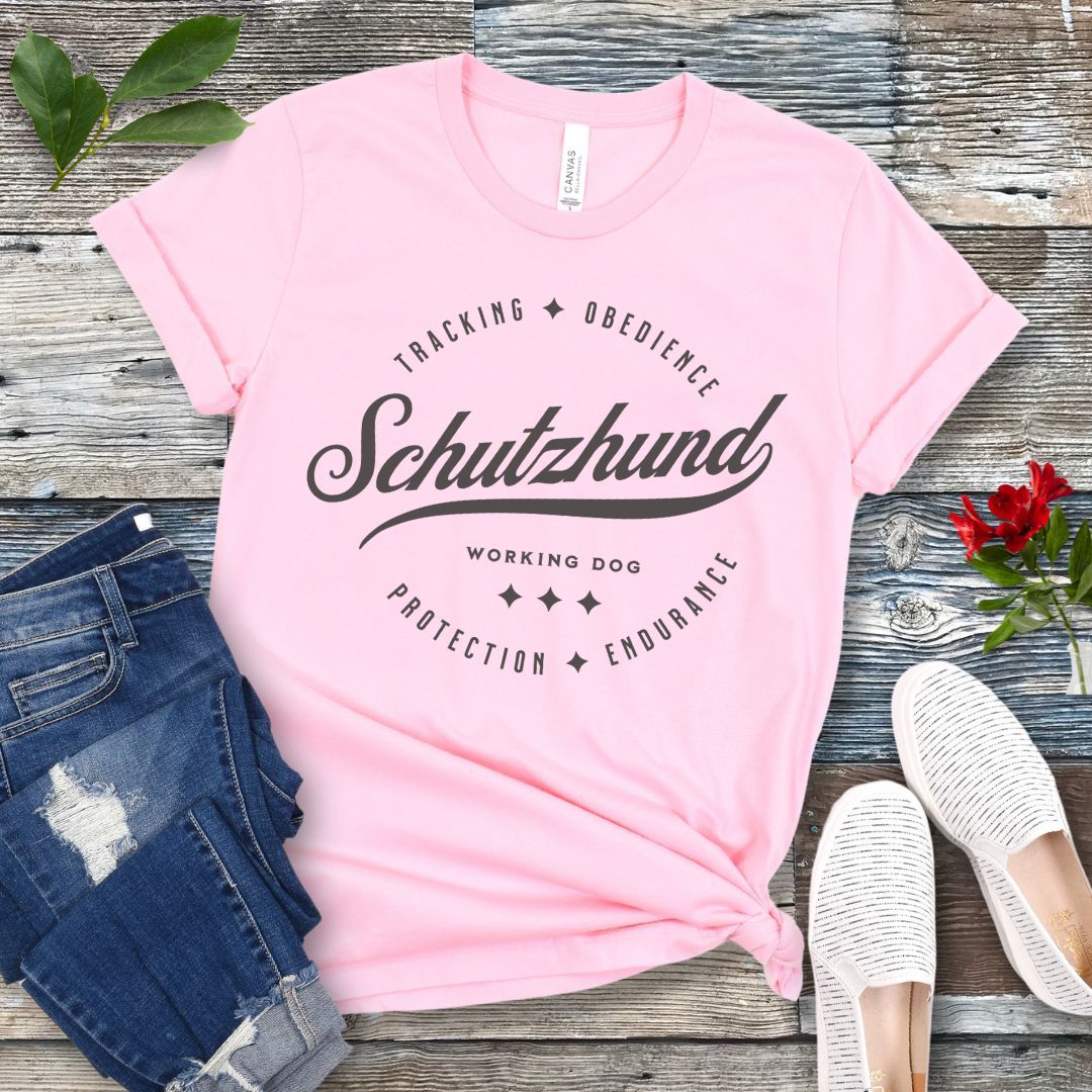A pink t-shirt with 'Schutzhund Working Dog' text and associated imagery in a vintage badge style design, displayed flat with a pair of blue jeans and white shoes.