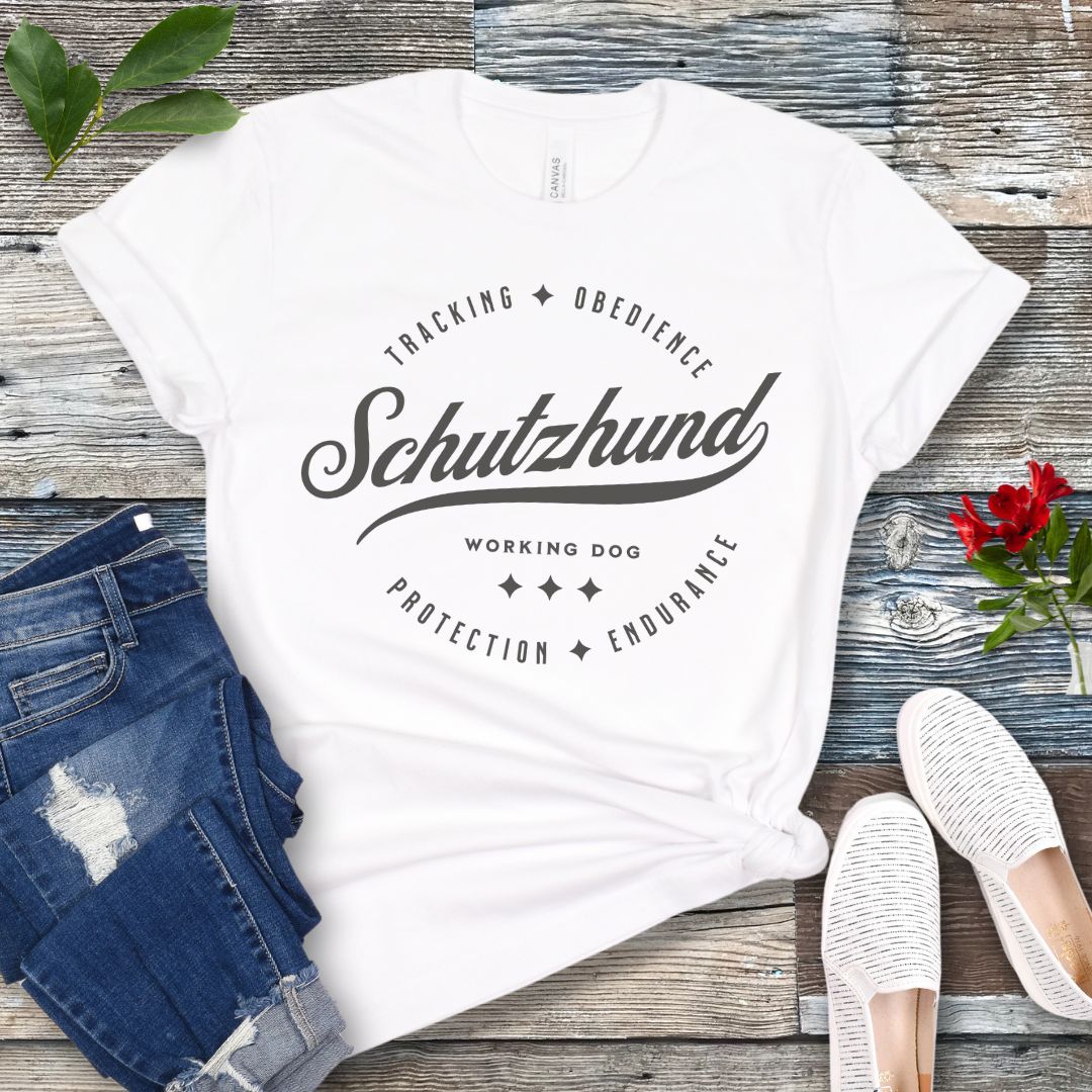 A white t-shirt with 'Schutzhund Working Dog' text and associated imagery in a vintage badge style design, displayed flat with a pair of blue jeans and white shoes.