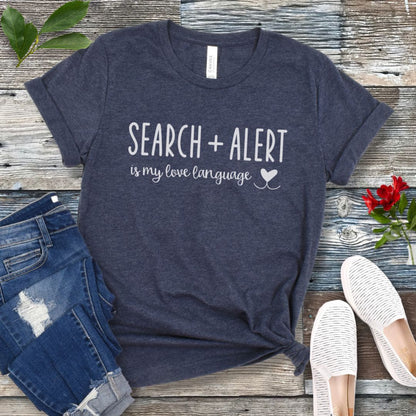 Heather navy blue canine scent work t-shirt with 'Search + Alert is my love language' text on a wooden surface with jeans and shoes.